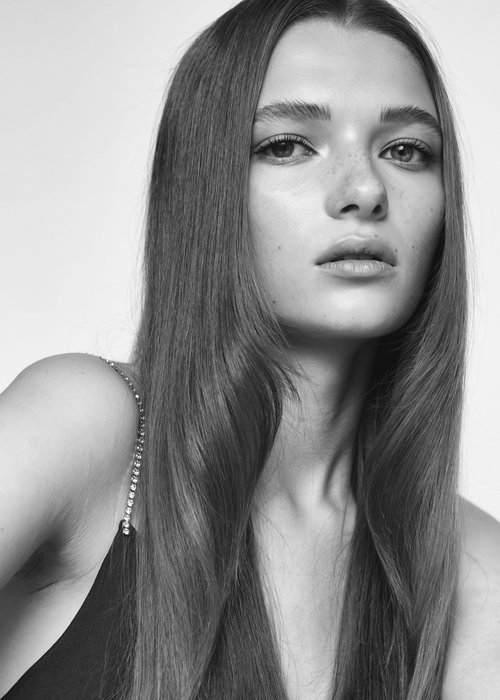 Close-up portrait of a young woman with long straight hair and subtle freckles wearing a thin-strapped dark top.