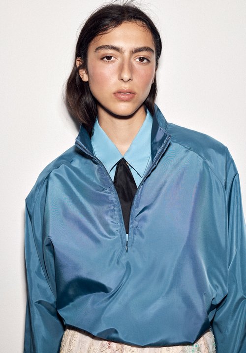 Close-up portrait of a woman wearing a blue zip jacket over a light blue shirt and black tie against a white background.
