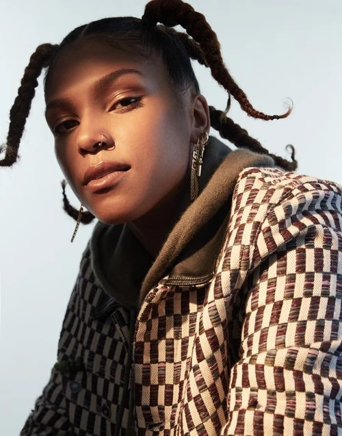 Young woman with unique hairstyle poses in a brown and beige checkered jacket against a soft blue backdrop in an eye-level portrait.