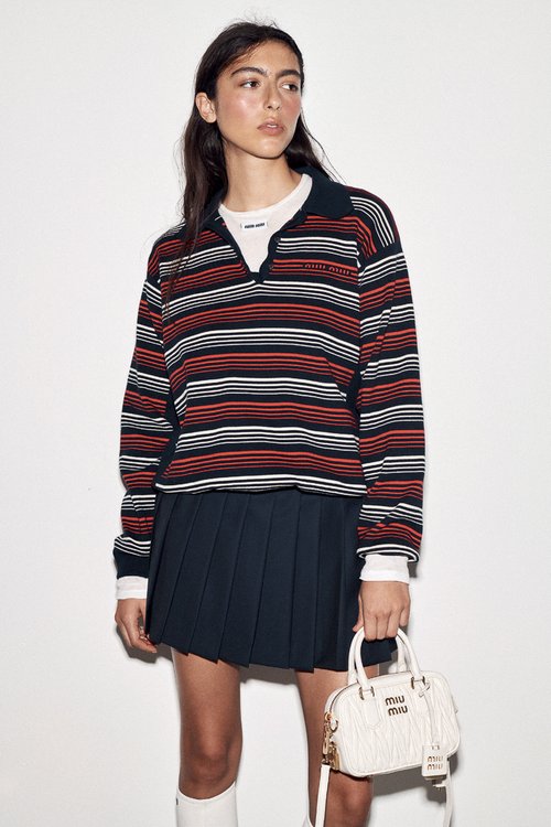 Model wearing striped polo shirt and pleated dark skirt holding white handbag against plain background
