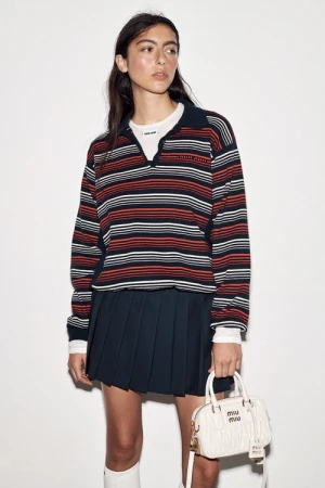 Model wearing striped polo shirt and pleated dark skirt holding white handbag against plain background
