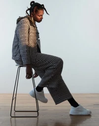 Model seated on a wire stool wearing a patterned black and white jacket and trousers with white sneakers
