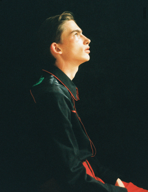Profile view of a young adult in a black jacket with red trim, looking upwards in a dark, underexposed indoor environment.