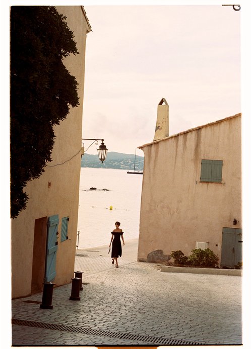 Woman in black dress walks on a cobblestone street between buildings toward beach and ocean under a cloudy sky in the daytime.