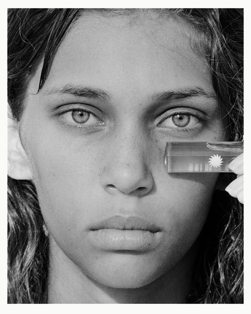 Monochrome portrait of a woman with light skin holding a clear tube of skincare product in front of her face in a beauty shot.