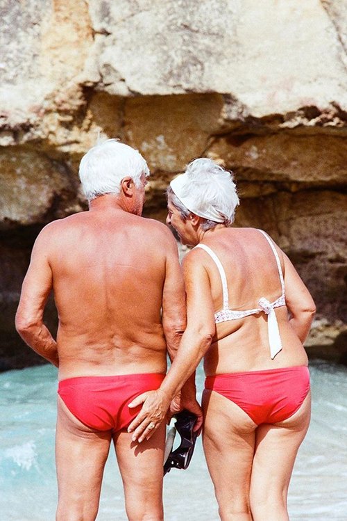 Older couple in red swimsuits standing on a beach with blue water and rock formations in background. Woman touches man's backside.