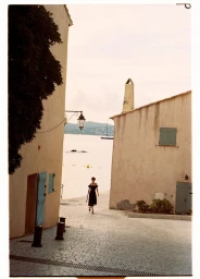 A woman in a dark off-the-shoulder dress walks down a cobbled alleyway toward a bright coastal view, captured by Ryan Brabazon.