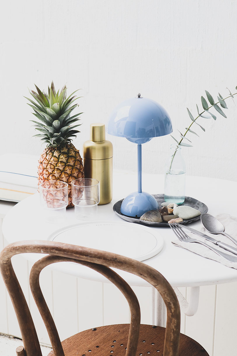 Still life on a table featuring a pineapple, bar shaker, glasses, a blue mushroom lamp, shells, and a chair in a bright indoor setting.
