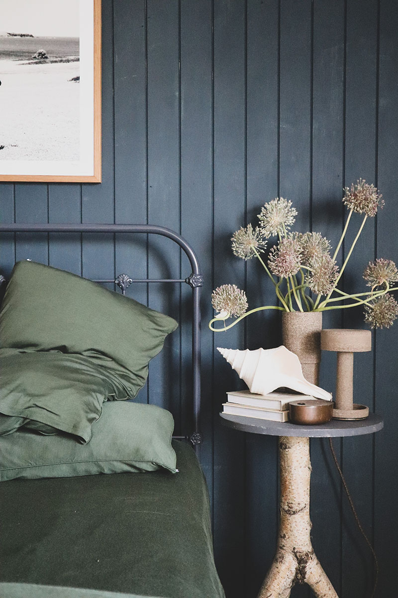 Bedroom interior with a bed featuring green linens, a nightstand, floral arrangement, and a framed picture on a blue panelled wall.