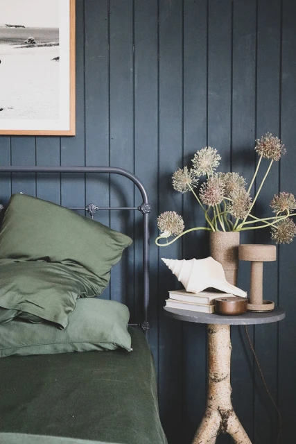 Bedroom interior with a bed featuring green linens, a nightstand, floral arrangement, and a framed picture on a blue panelled wall.