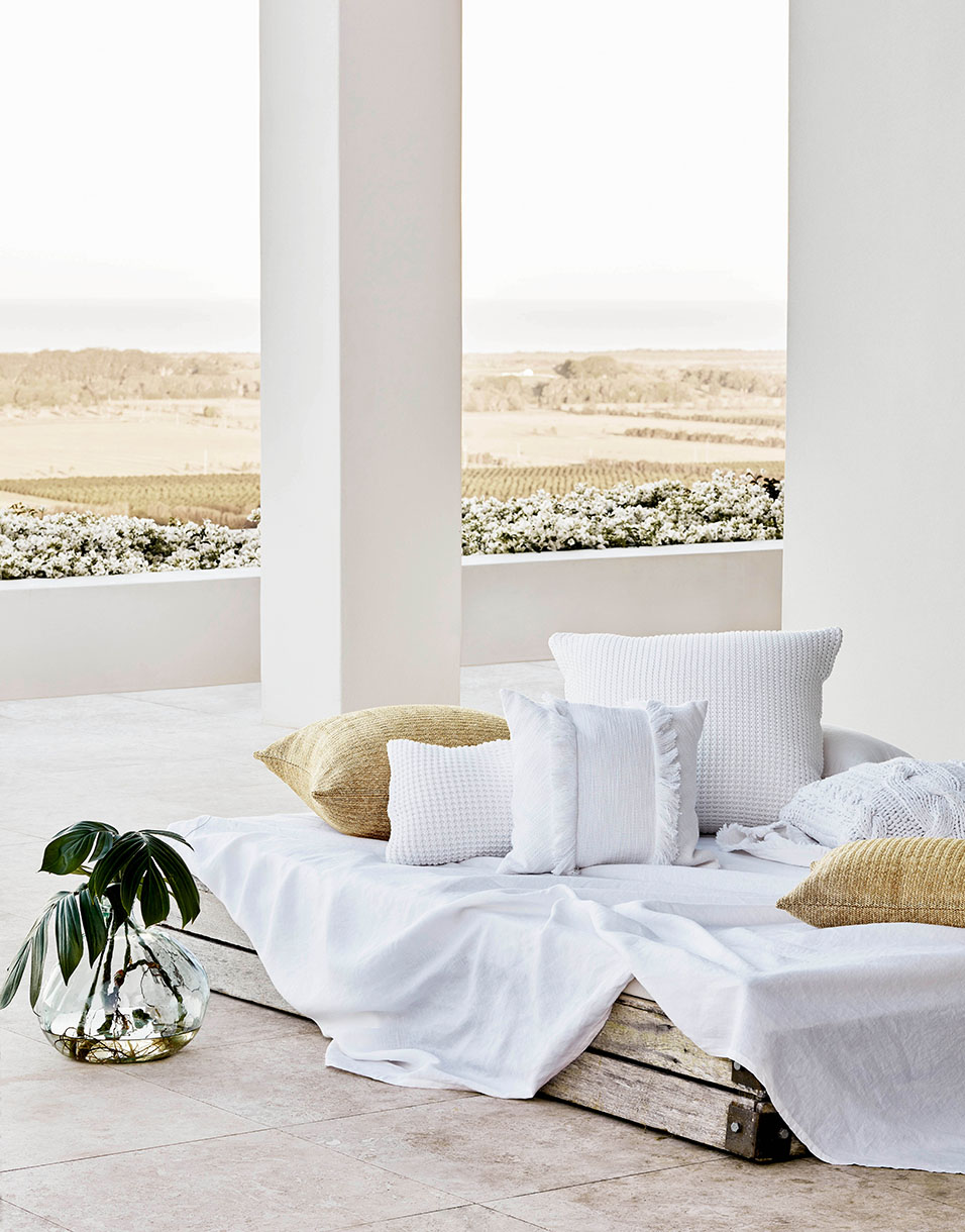 Outdoor patio setup with white cushions and throw blanket, complemented by a glass vase with green leaves and a neutral landscape view.