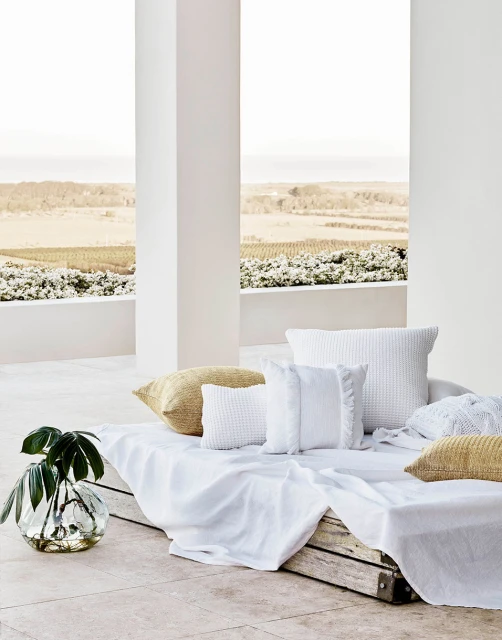 Outdoor patio setup with white cushions and throw blanket, complemented by a glass vase with green leaves and a neutral landscape view.