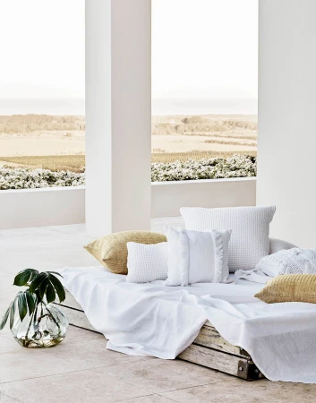 Outdoor patio setup with white cushions and throw blanket, complemented by a glass vase with green leaves and a neutral landscape view.