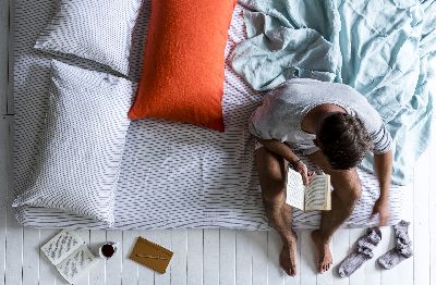 Overhead shot of a man reading a book in bed surrounded by pillows, a cup, and socks on a white wooden floor.