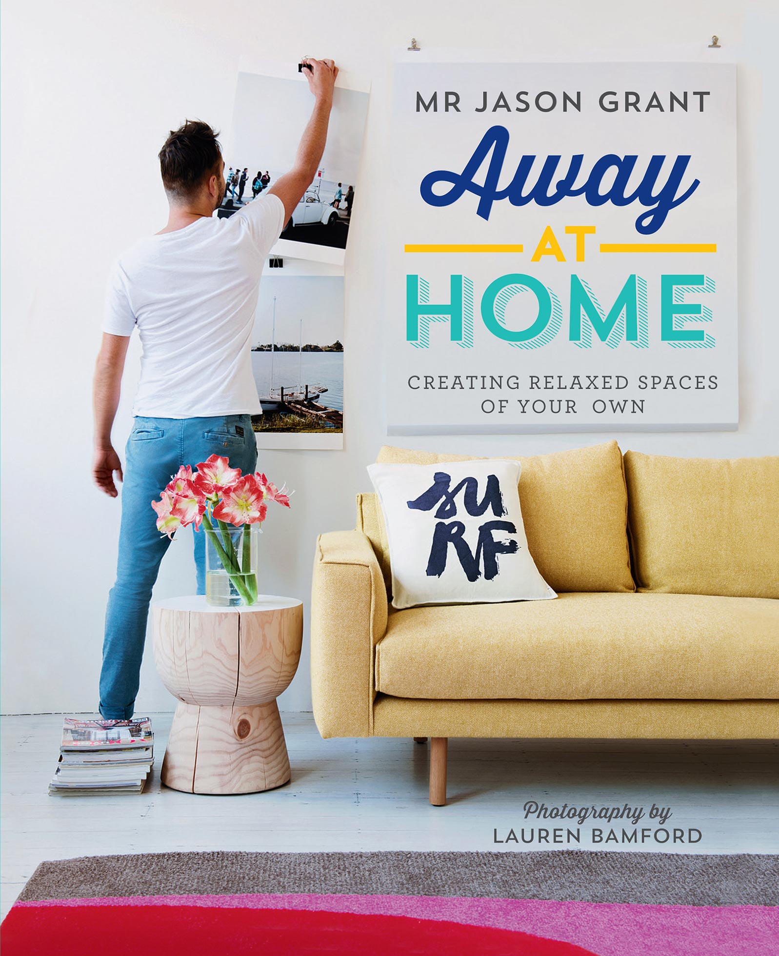 Person hanging poster on wall next to book cover of Away at Home by Mr Jason Grant, with a yellow couch and floral arrangement.