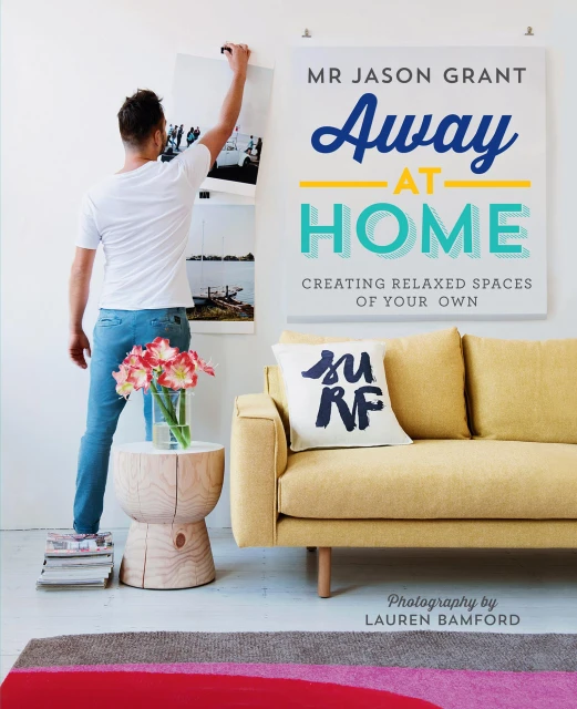 Person hanging poster on wall next to book cover of Away at Home by Mr Jason Grant, with a yellow couch and floral arrangement.