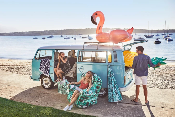 A turquoise vintage VW bus parked near a beach with two women relaxing while a man carries beach gear near the vehicle.