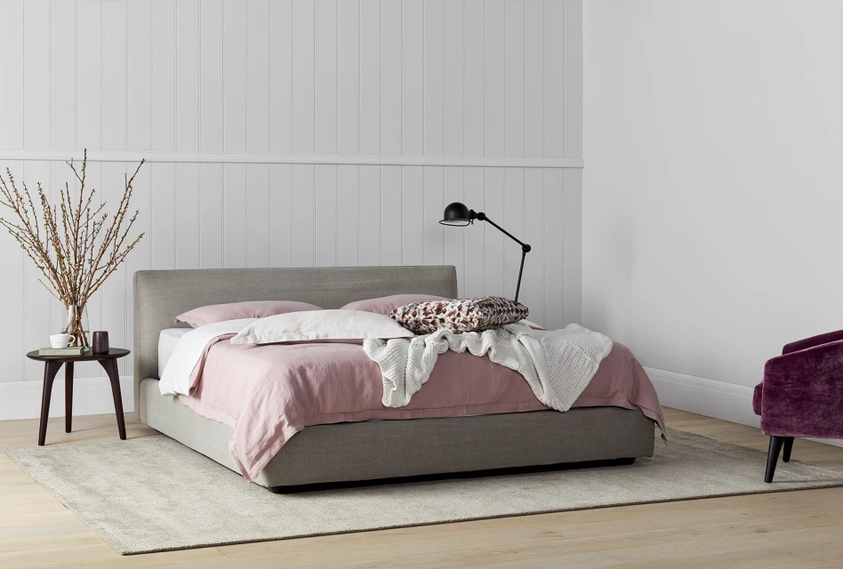 Bedroom interior featuring a made bed with pink linen, a side table with vase, a black lamp, and a purple velvet armchair.