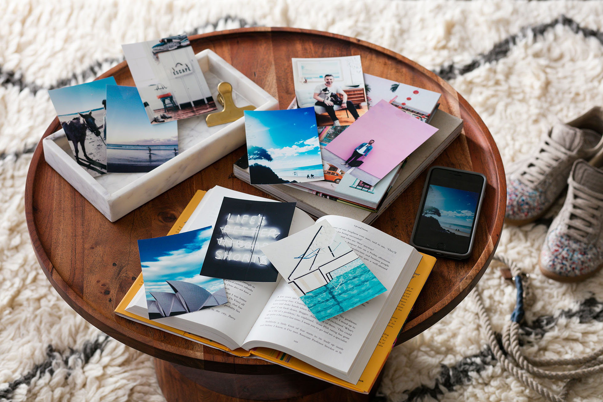 An overhead shot shows an open book, a cell phone, photographs, and shoes on a round wood table with a white patterned rug.