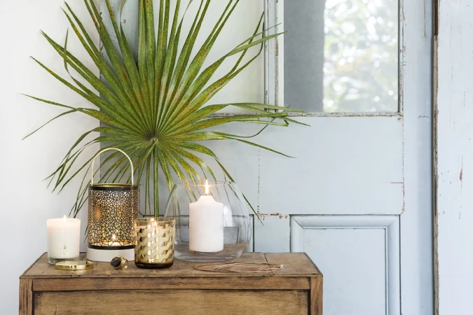 Still life composition of candles, golden lantern, jewelry, and palm leaf on a wooden table against a faded blue door backdrop.