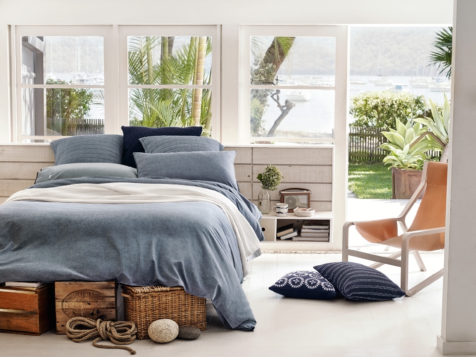 Bedroom interior with a denim blue duvet on a platform bed, wooden crates, nautical rope, and natural light from windows overlooking a bay.