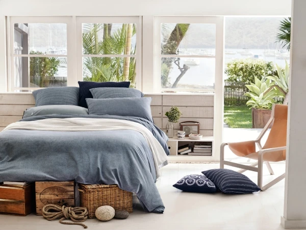 Bedroom interior with a denim blue duvet on a platform bed, wooden crates, nautical rope, and natural light from windows overlooking a bay.