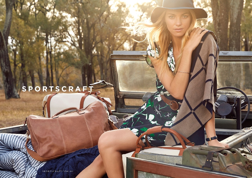 A blonde woman in a floral dress and hat sits atop a vintage vehicle in a sunlit outdoor setting, surrounded by luggage and rustic charm.
