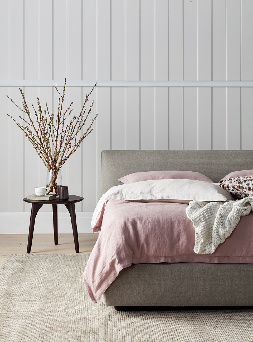 Bedroom interior featuring a gray upholstered bed with pink linen, a round side table with a vase of branches, and a light-colored rug.