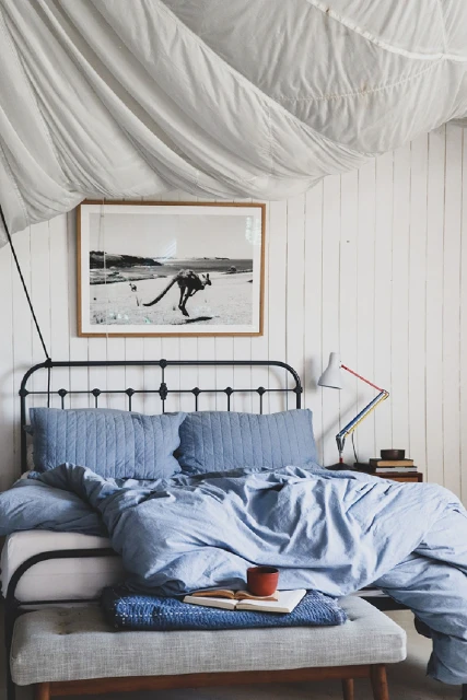 Bedroom interior with a metal bed frame, blue bedding, a bench, and a framed photograph on the white wood panelled wall above the bed.
