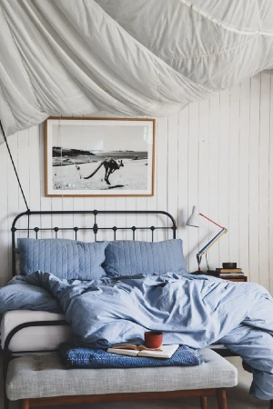 Bedroom interior with a metal bed frame, blue bedding, a bench, and a framed photograph on the white wood panelled wall above the bed.