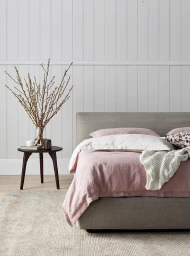 A made bed with pink linen bedding, white pillows, and a knitted throw next to a dark wood side table with a vase of bare branches.