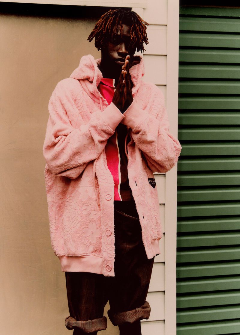 An adult male model with dark skin poses in a pink sherpa hoodie, reddish top, and brown pants against an indoor wall.