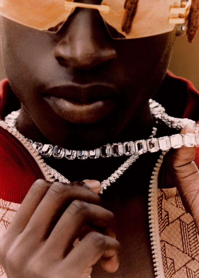 Close-up of a dark-skinned adult wearing orange sunglasses and layered diamond necklaces, posing indoors with a red and brown patterned jacket.