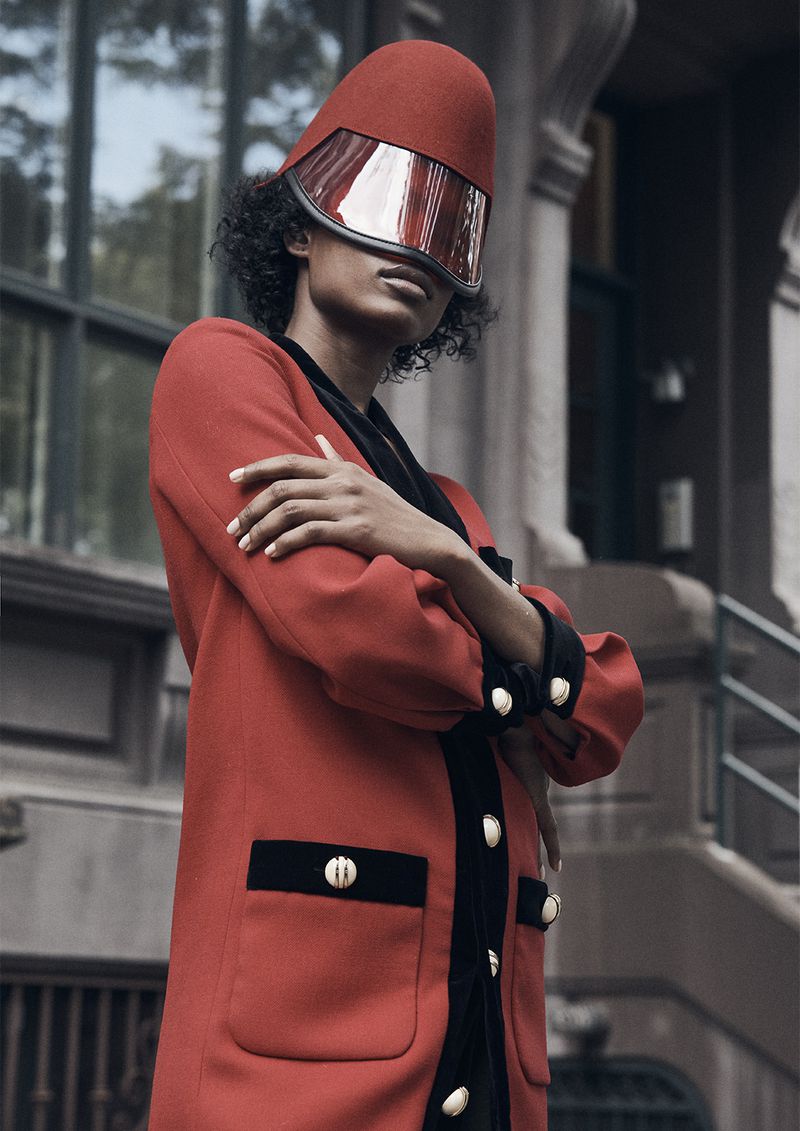 Fashionable woman poses outdoors in a red coat with black trim and a red visor hat. Urban setting with natural light.