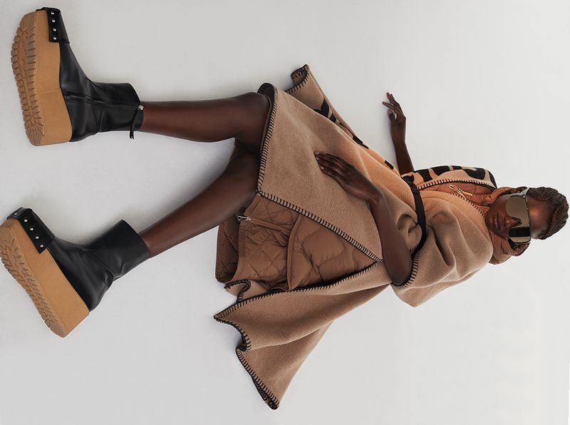 Fashion shot of a Black female model wearing a tan wrap coat and black platform boots lying on a white studio surface.