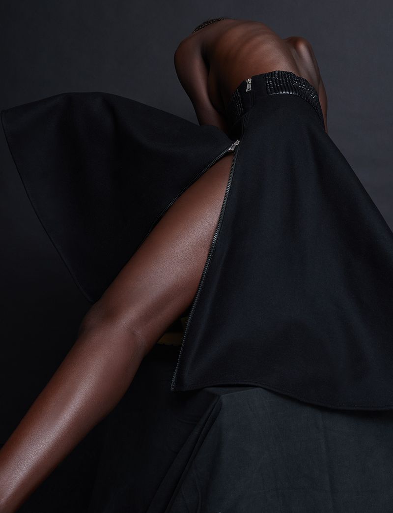 Dark-skinned human figure with a black slit skirt against a dark backdrop, accentuating shape and texture through studio lighting.