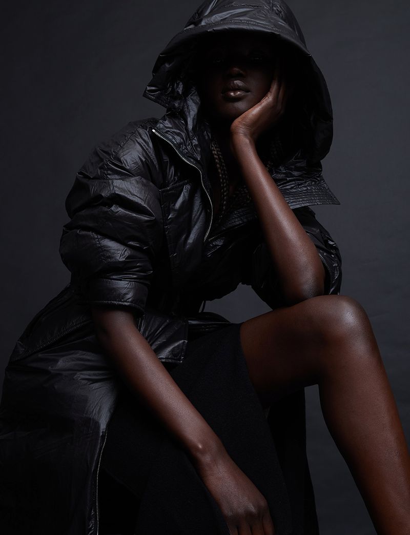 Dark-skinned model in a hooded, black puffer coat sitting in a studio with a thoughtful pose and atmospheric lighting.