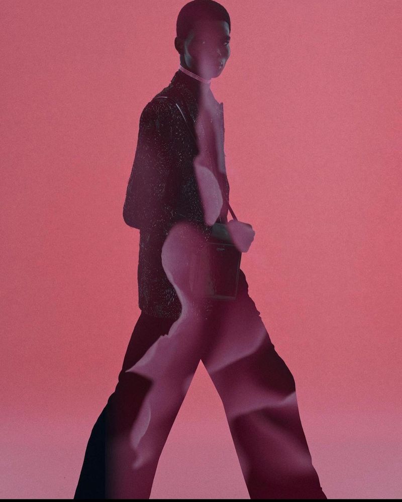 A slender, dark-skinned man in a dark suit, walking with a bag, set against a soft pink backdrop and unique artistic shadow effects.