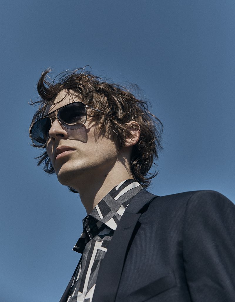 Adult male fashion portrait under blue sky. Man with brown hair wears sunglasses, patterned shirt, and blazer in an outdoor setting.