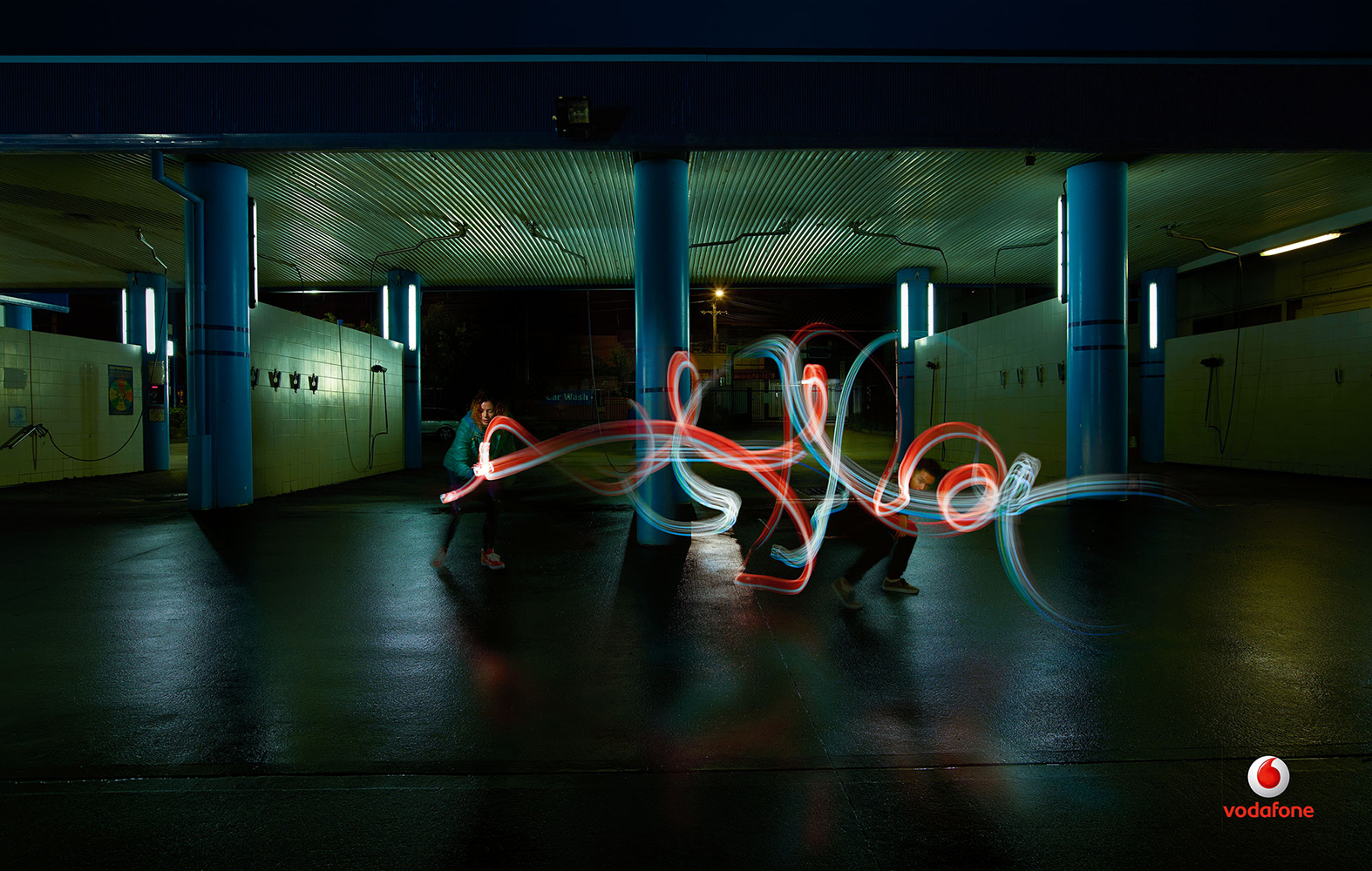 Long exposure shot of two figures using light trails to create abstract patterns under a structure with blue support pillars.