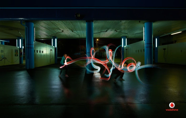 Long exposure shot of two figures using light trails to create abstract patterns under a structure with blue support pillars.