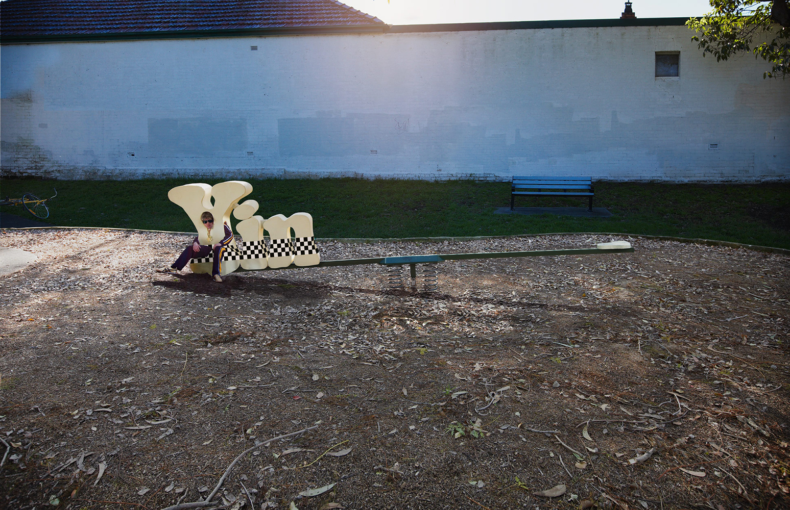 Adult female in tracksuit poses on seesaw at playground on sunny day with white brick wall and greenery backdrop in wide outdoor shot.