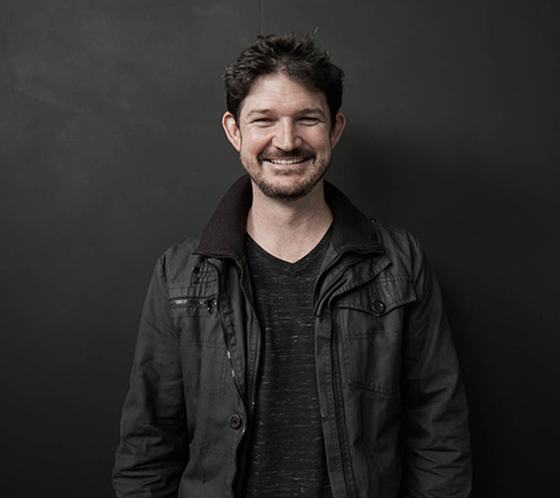 Portrait of a smiling adult male wearing a dark jacket and t-shirt, set against a dark grey background, medium shot, eye-level angle.