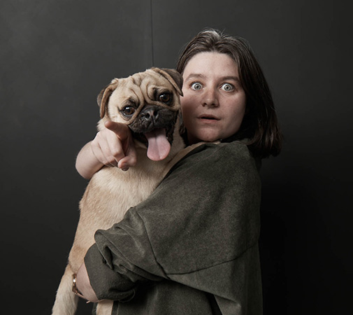 Woman embraces a light-brown pug dog with tongue out in a studio setting with black background; the woman points her index finger.