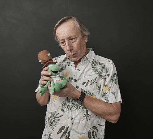 Man holding plush toy against dark background, wearing a Hawaiian shirt and a smartwatch, looking down with slight smile.