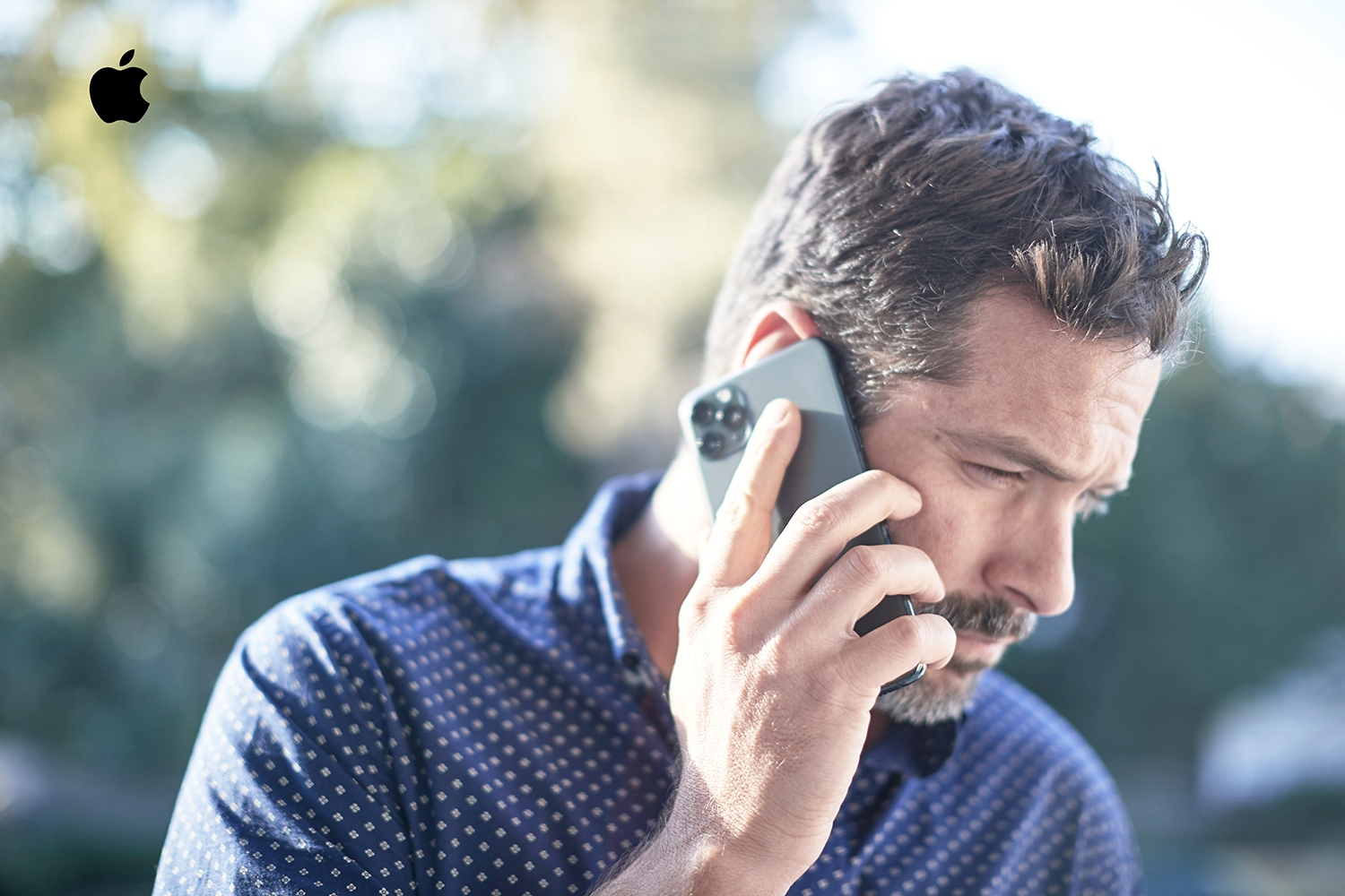 Man in blue patterned shirt on phone outdoors with Apple logo. Man with beard on mobile call, blurred green background, sunny.