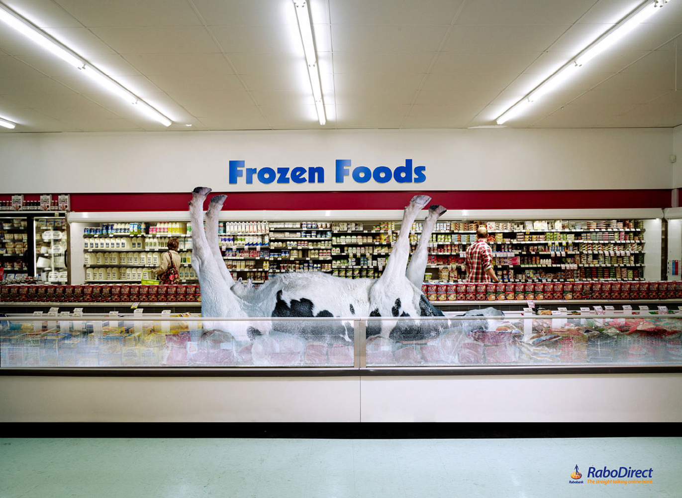 A cow lies belly-up inside a supermarket freezer case, surrounded by frozen foods, with a customer shopping in the background.