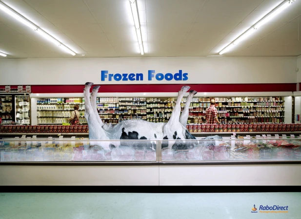 A cow lies belly-up inside a supermarket freezer case, surrounded by frozen foods, with a customer shopping in the background.
