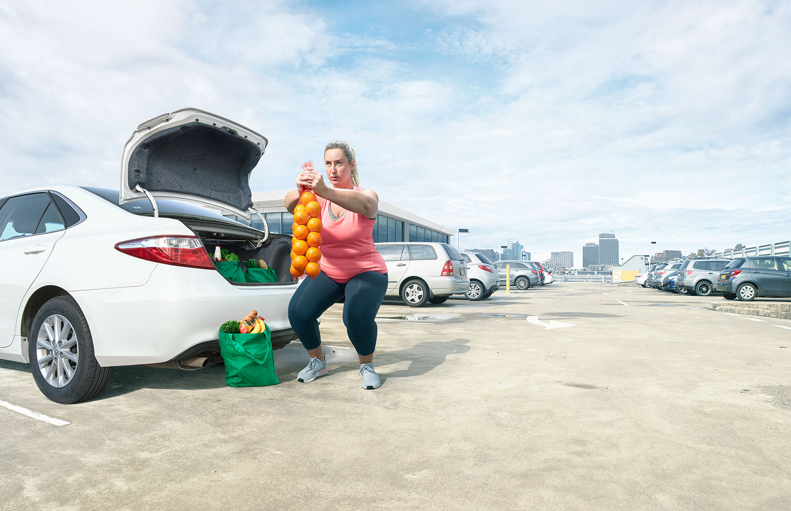 Active woman squats with bag of oranges near car in parking lot. Fitness combined with healthy grocery shopping and automotive transport.