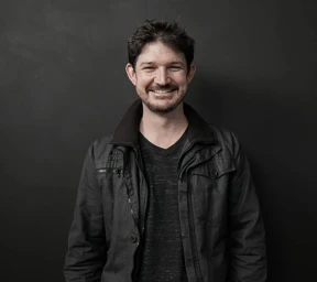 Portrait of a smiling man wearing a dark jacket against a dark background, photographed in Sydney by Andreas Bomert.